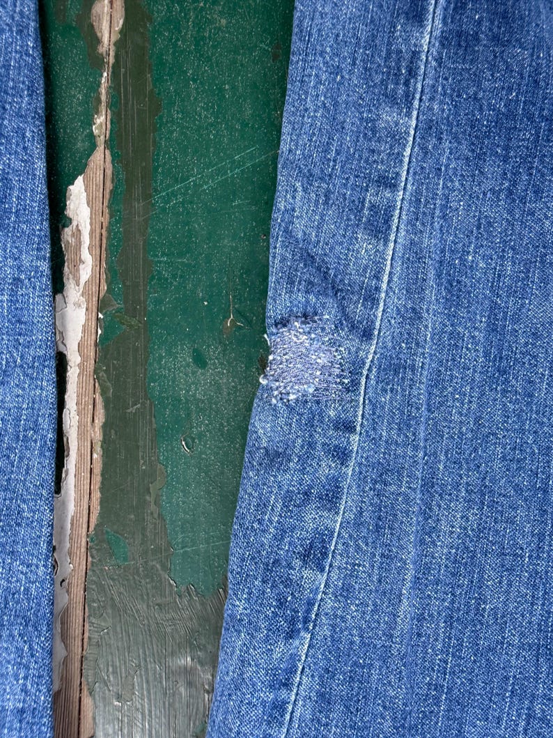May include: Close-up of blue denim jeans with a distressed patch. The jeans show signs of wear and tear, with a visible hole and frayed edges. The background is a weathered, green-painted wooden surface.
