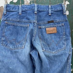 May include: Close-up of the back of blue denim jeans. The jeans have two back pockets with decorative stitching and a brown leather patch with the brand name "Wrangler". The jeans have a classic, vintage style.