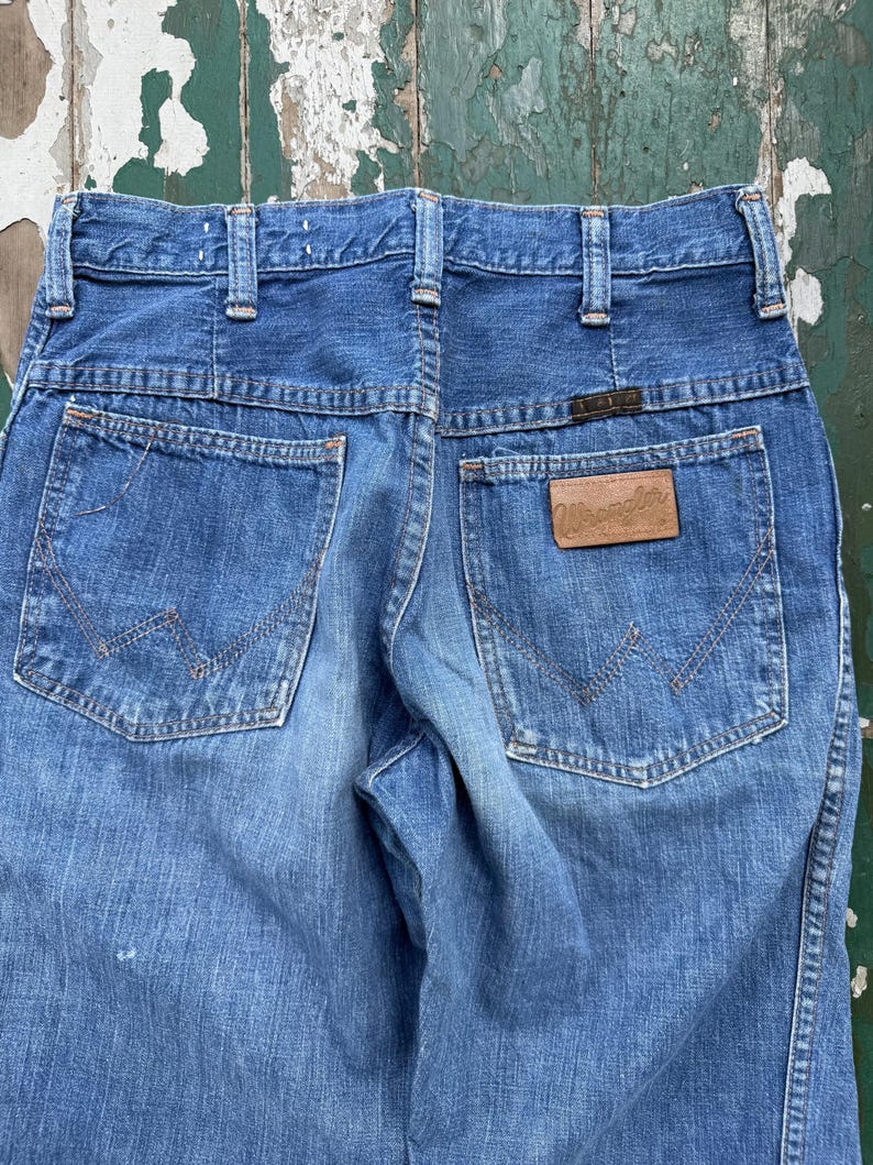 May include: Close-up of the back of blue denim jeans. The jeans have two back pockets with decorative stitching and a brown leather patch with the brand name "Wrangler". The jeans have a classic, vintage style.