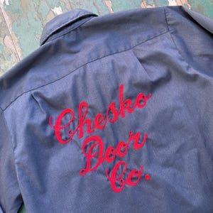 May include: A blue work shirt with a collar and long sleeves. The back of the shirt features the embroidered text "Chesko Door Co." in red script. The shirt is made of a durable fabric.