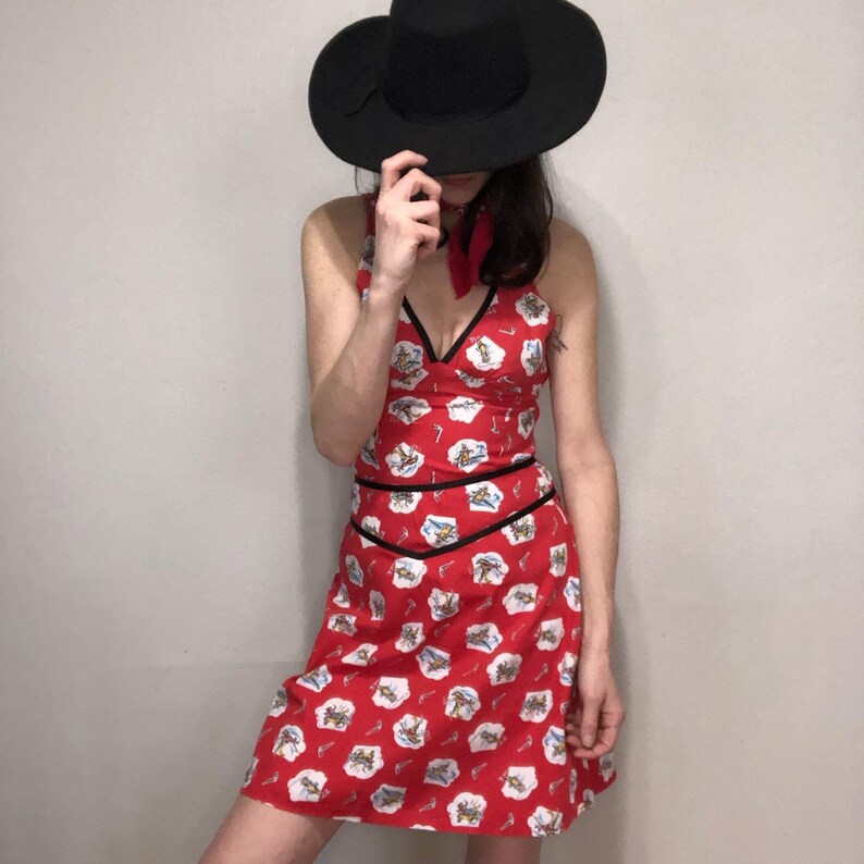 Peut inclure: Une robe rouge avec une bordure noire et un imprim&eacute; blanc de cowboys et de chevaux. La robe a un d&eacute;collet&eacute; halter et un corsage ajust&eacute;. Le mannequin porte un chapeau noir.