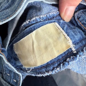 May include: Close-up of blue denim jeans, showing the inside waistband and a blank, rectangular tag. The denim fabric has a classic, worn appearance with visible stitching and a light-colored tag.