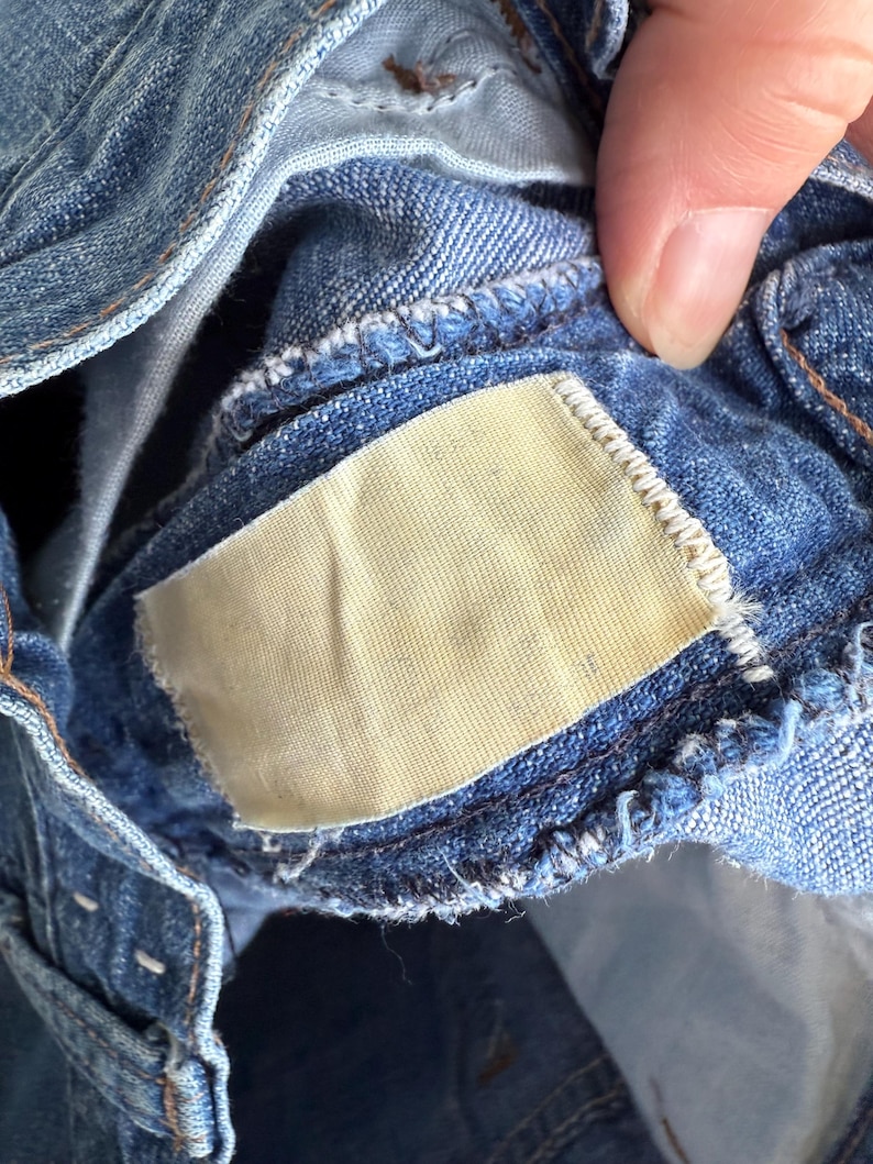 May include: Close-up of blue denim jeans, showing the inside waistband and a blank, rectangular tag. The denim fabric has a classic, worn appearance with visible stitching and a light-colored tag.