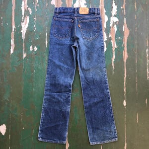 May include: Blue denim jeans with a classic five-pocket design. The jeans feature orange stitching, a brown leather patch, and a faded appearance. The jeans are laid flat, showcasing the back pockets and the Levi's brand label.