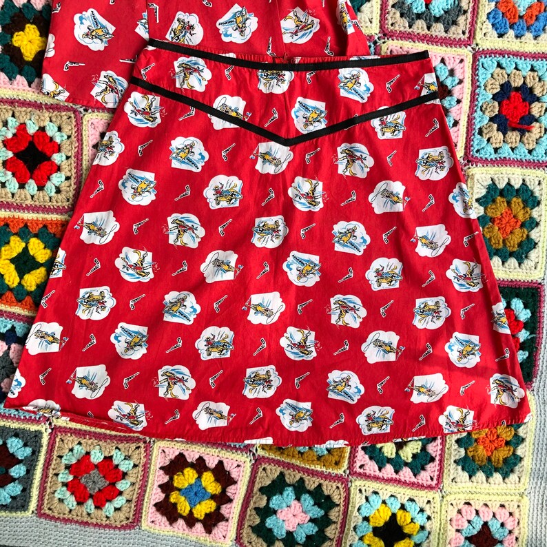 Peut inclure: Une jupe rouge avec une bordure noire et un motif de petits avions. La jupe est faite d'un tissu l&eacute;ger et a une forme ample et &eacute;vas&eacute;e.