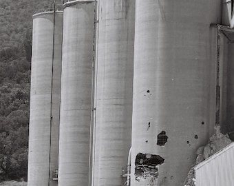 May include: Four large, gray concrete silos stand side-by-side. One silo has a large hole in the side, revealing the interior.