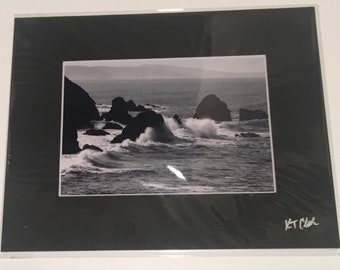 May include: Black and white photograph of a rocky coastline with crashing waves. The photo is framed with a black mat and signed "KT Clark" in the bottom right corner.