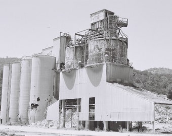 May include: A black and white photo of an abandoned industrial building with large silos and a tall structure with a platform on top.