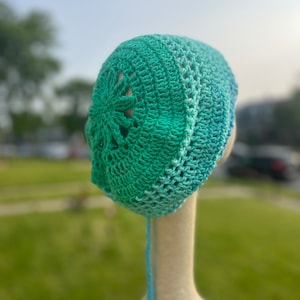 May include: A crocheted hat with a blue and green ombre colour scheme. The hat has a large openwork design on the top and a long tie.