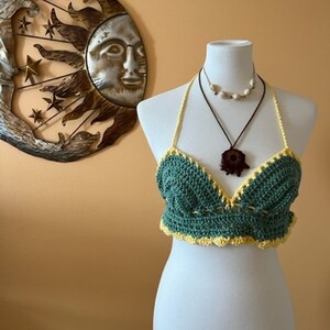 May include: A handmade green and yellow crochet top displayed on a mannequin. The halter top features a scalloped edge and a decorative design. Two necklaces are worn on the mannequin. A metal sun and star wall hanging is in the background.