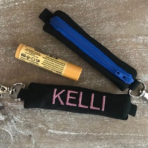Puede incluir: Soportes de bálsamo labial de tela negra con cierres plateados. Uno tiene una cremallera azul y el otro el nombre "KELLI" bordado en rosa. También hay un tubo de bálsamo labial.