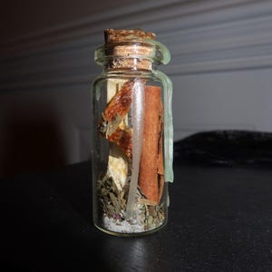 May include: A small clear glass bottle sealed with a cork and light green wax. Inside the bottle are dried herbs, a cinnamon stick, and a piece of dried orange peel. The contents are layered with a fine white granular substance at the bottom.