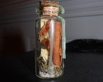 May include: A small clear glass bottle sealed with a cork and light green wax. Inside the bottle are dried herbs, a cinnamon stick, and a piece of dried orange peel. The contents are layered with a fine white granular substance at the bottom.