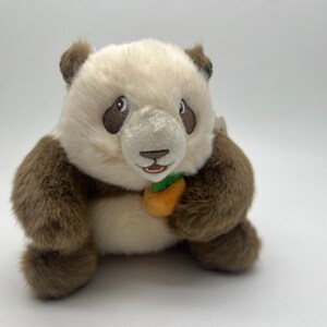 May include: A plush panda bear with a cream-colored face and belly, brown fur on its limbs and ears, and black eyes. The panda is holding a small orange carrot with green leaves.