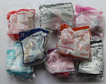 Puede incluir: Un conjunto de juguetes sellados de McDonald's Happy Meal con Snoopy de la tira cómica Peanuts. Los juguetes están en bolsas de plástico transparente con gráficos coloridos y texto, incluyendo "Snoopy's World" e ilustraciones de Snoopy.