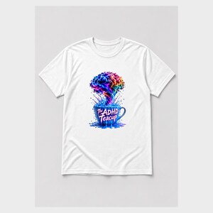 Puede incluir: Una camiseta blanca de manga corta con un gr&aacute;fico de una taza de t&eacute; azul de la que sale una nube colorida y arremolinada de humo o vapor. El texto "The ADHD Teacup" est&aacute; impreso con una fuente estilizada en la parte frontal de la taza.