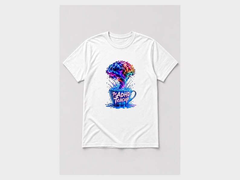 Puede incluir: Una camiseta blanca de manga corta con un gr&aacute;fico de una taza de t&eacute; azul de la que sale una nube colorida y arremolinada de humo o vapor. El texto "The ADHD Teacup" est&aacute; impreso con una fuente estilizada en la parte frontal de la taza.