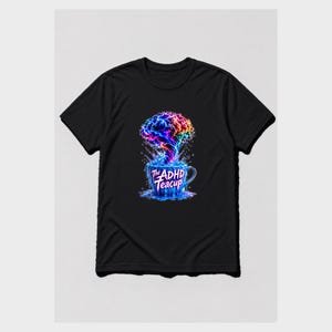 Puede incluir: Una camiseta negra con un gr&aacute;fico de una taza de t&eacute; azul que rebosa con una nube multicolor en forma de cerebro. El texto "The ADHD Teacup" est&aacute; impreso en blanco en la parte frontal de la taza, rodeado de salpicaduras de l&iacute;quido azul.