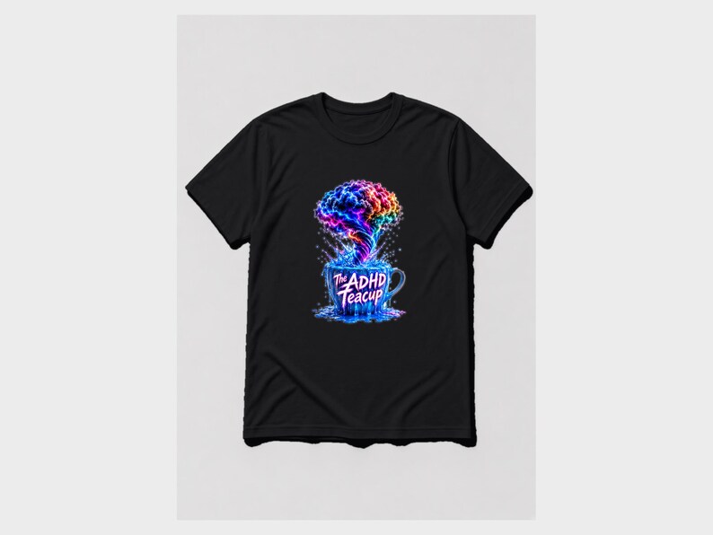 Puede incluir: Una camiseta negra con un gr&aacute;fico de una taza de t&eacute; azul que rebosa con una nube multicolor en forma de cerebro. El texto "The ADHD Teacup" est&aacute; impreso en blanco en la parte frontal de la taza, rodeado de salpicaduras de l&iacute;quido azul.