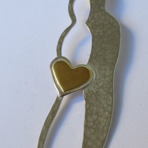 May include: A silver necklace with a pendant shaped like two figures holding a gold heart. The pendant has a textured surface.