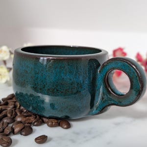 May include: A ceramic mug with a deep teal and dark brown mottled glaze. The mug features a wide, rounded body and a prominent, circular handle. The exterior surface has a glossy finish with visible color variations throughout the piece.