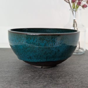 May include: A round ceramic bowl featuring a deep teal and blue mottled glaze on the exterior and interior, with a dark brown, unglazed base. The bowl has a smooth, curved rim and a wide opening, showcasing a rich, variegated color finish.