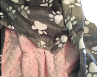 May include: A soft, gray and pink blanket with a paw print pattern. One side of the blanket is gray with white paw prints, and the other side is pink with a textured pattern.