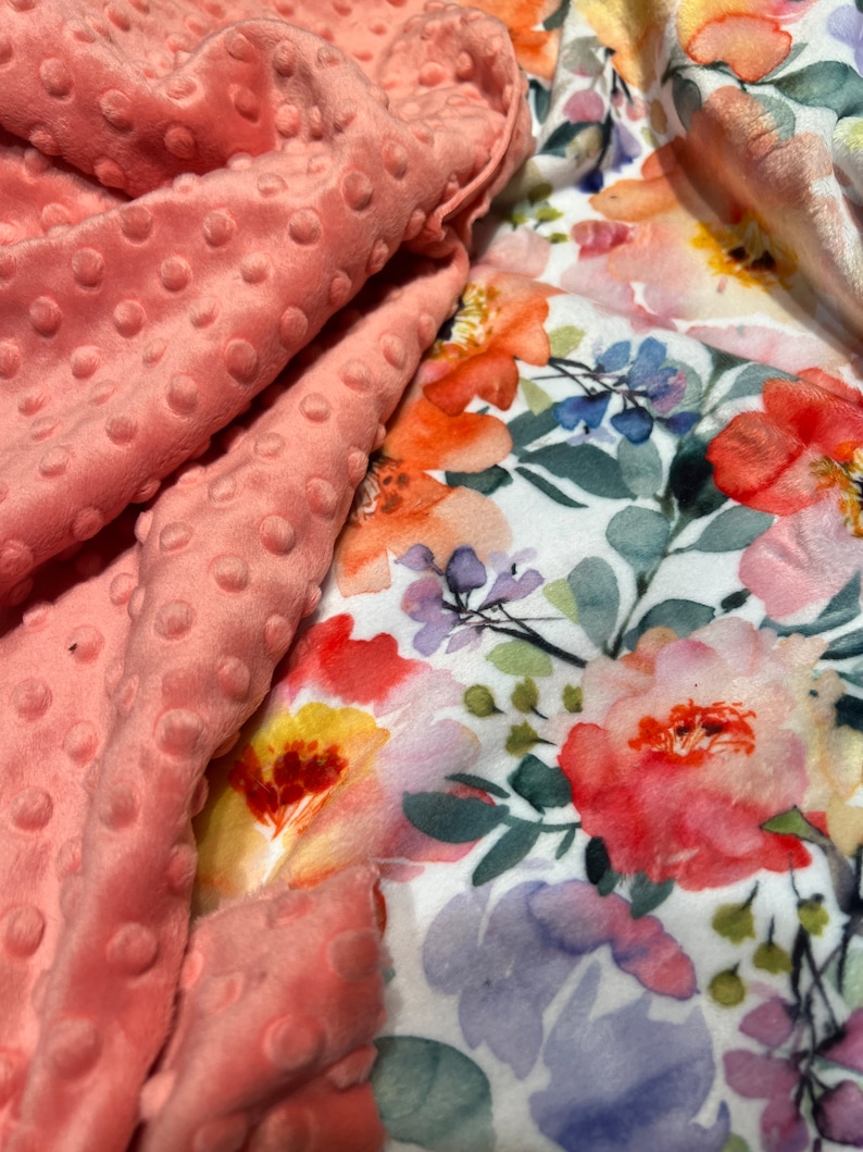 May include: A pink minky fabric with a white background featuring a watercolor floral print. The floral print includes pink, orange, yellow, and purple flowers.