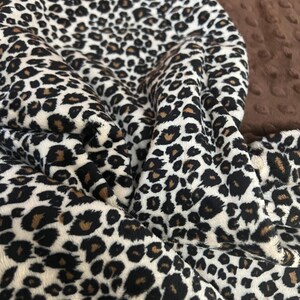 May include: A close-up of a white, brown, and black leopard print fabric. The fabric is soft and plush, with a fuzzy texture.