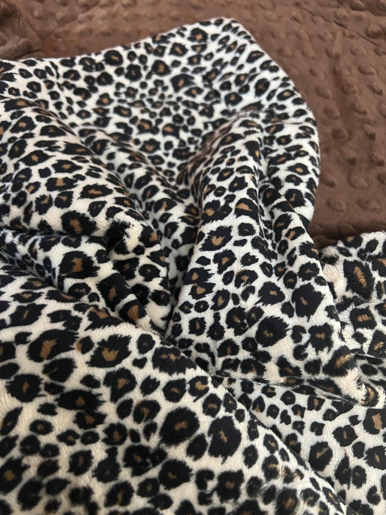 May include: A close-up of a white, brown, and black leopard print fabric. The fabric is soft and plush, with a fuzzy texture.