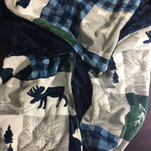May include: A cozy blanket featuring a patchwork design with moose and pine tree silhouettes. The blanket has a color scheme of navy blue, white, gray, and green. The pattern includes plaid and solid color blocks.