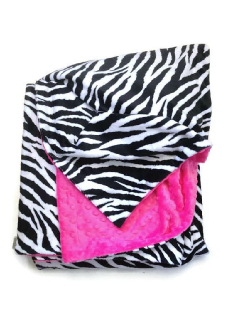 May include: A black and white zebra print blanket with a bright pink fleece lining. The blanket is folded in half and the pink side is facing up.