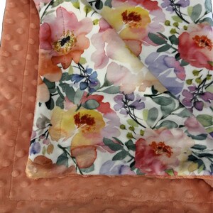 May include: A soft, peach-colored minky baby blanket with a white floral print. The blanket features a watercolor-style design with pink, yellow, and purple flowers.