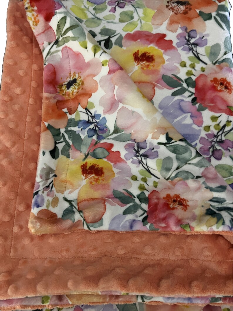 May include: A soft, peach-colored minky baby blanket with a white floral print. The blanket features a watercolor-style design with pink, yellow, and purple flowers.