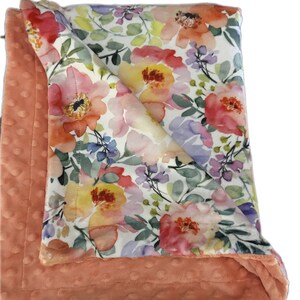 May include: A soft, peach-colored baby blanket with a floral print. The blanket is made of two layers, a soft, plush fabric on one side and a cotton fabric with a watercolor floral print on the other side.