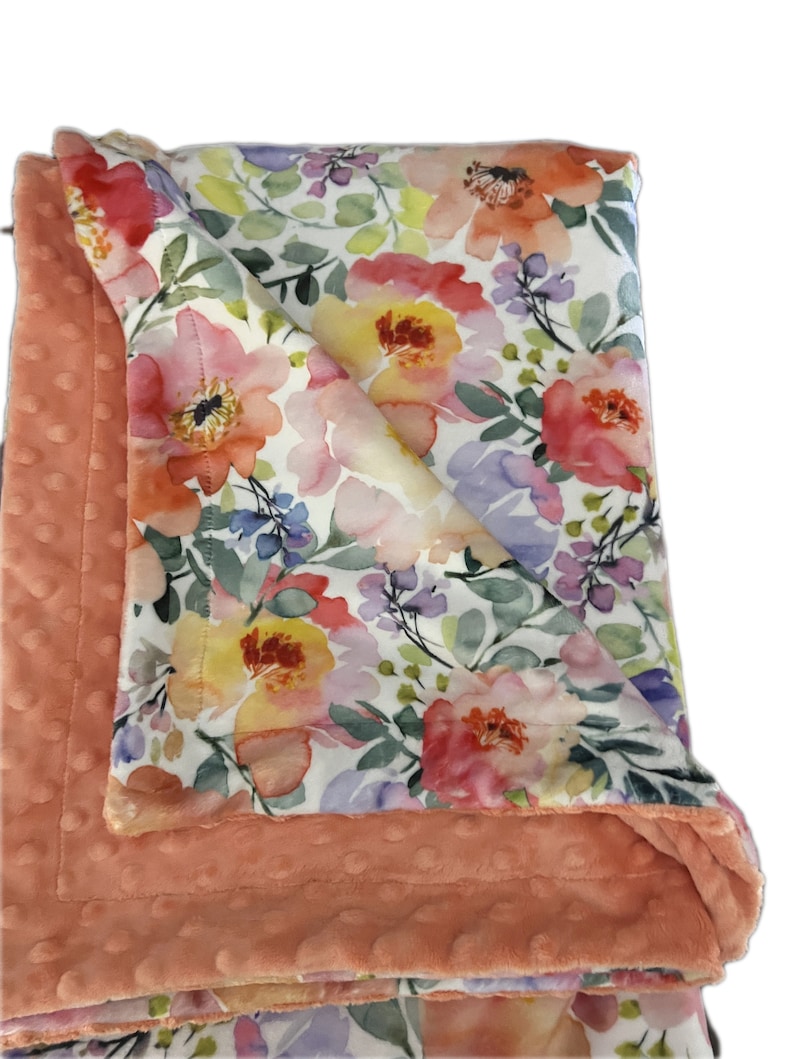 May include: A soft, peach-colored baby blanket with a floral print. The blanket is made of two layers, a soft, plush fabric on one side and a cotton fabric with a watercolor floral print on the other side.