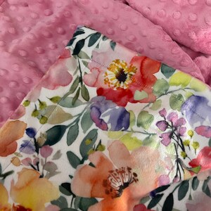 May include: A white fabric with a watercolor floral print featuring pink, yellow, orange, and purple flowers. The fabric is folded over a pink minky fabric with small raised dots.