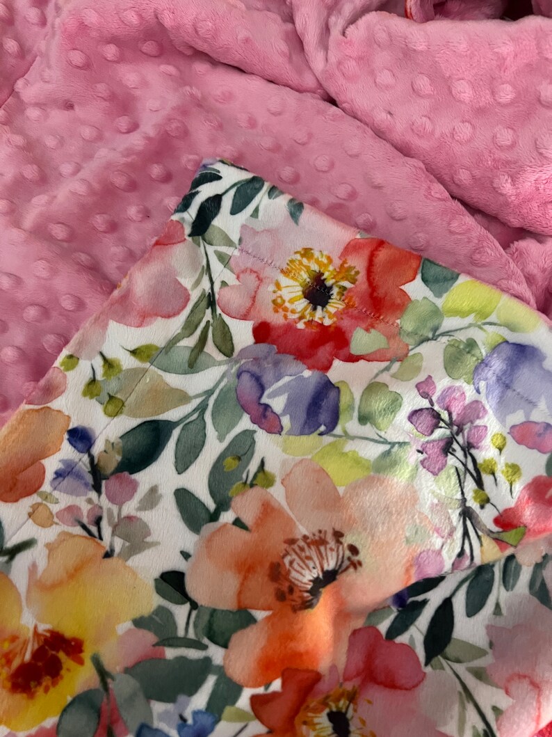 May include: A white fabric with a watercolor floral print featuring pink, yellow, orange, and purple flowers. The fabric is folded over a pink minky fabric with small raised dots.