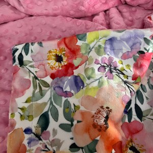 May include: A pink and white floral print fabric with a soft, plush pink minky backing. The floral print features a variety of flowers in shades of pink, orange, yellow, and purple.