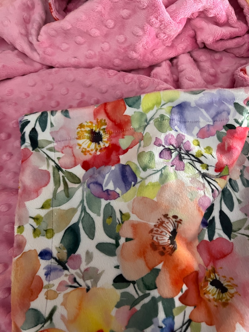 May include: A pink and white floral print fabric with a soft, plush pink minky backing. The floral print features a variety of flowers in shades of pink, orange, yellow, and purple.
