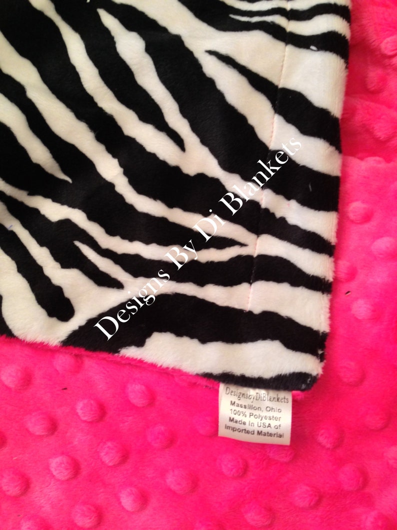 May include: A pink and black zebra print baby blanket with a minky dot fabric on the back. The blanket has a tag that reads "Designs By Di Blankets Massillon, Ohio 100% Polyester Made in USA of Imported Material."