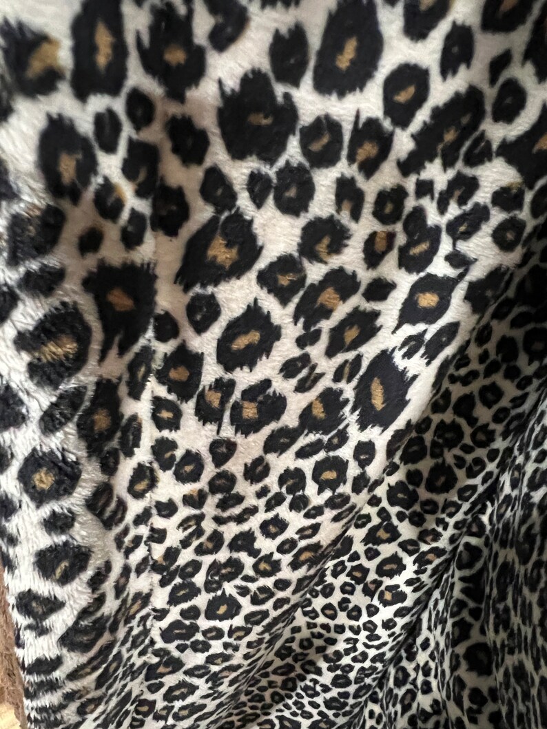 May include: Close-up of a white fabric with a black and brown leopard print pattern. The fabric is soft and plush, with a fuzzy texture.