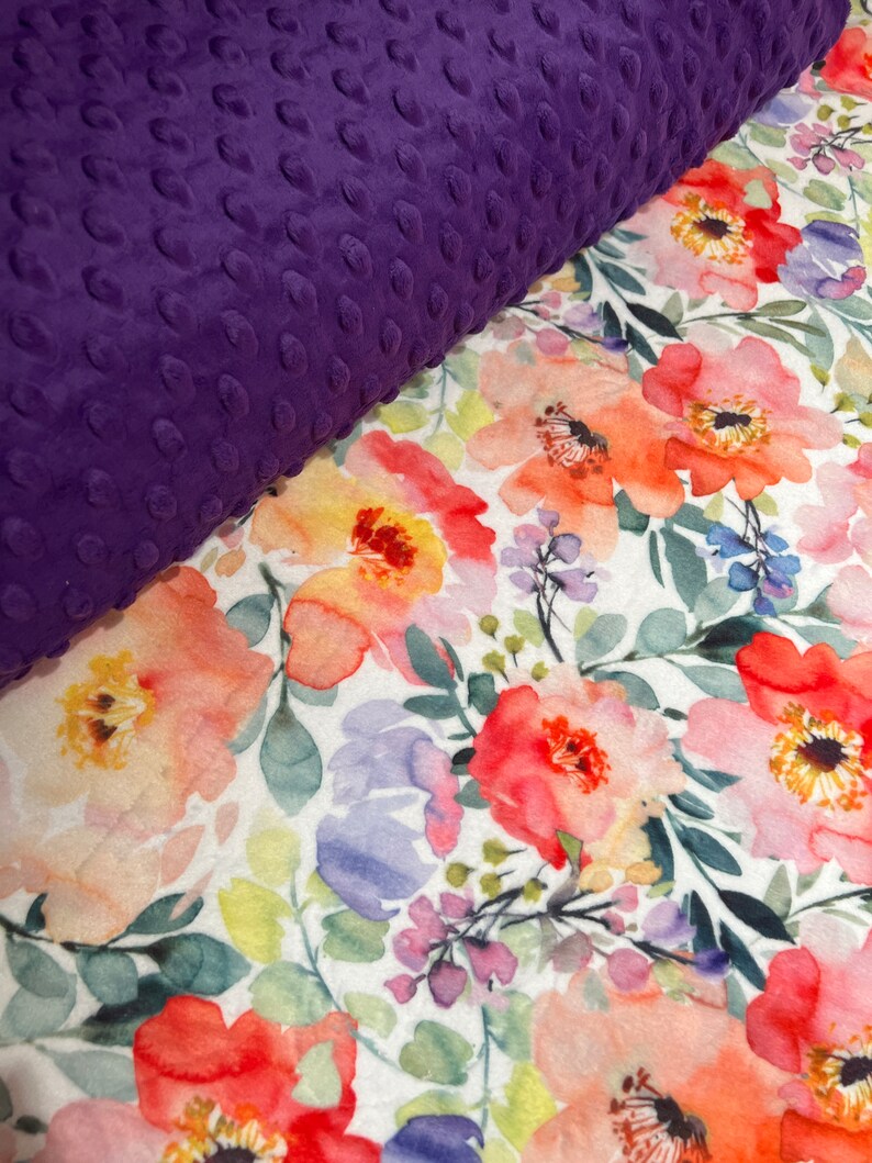 May include: A purple plush fabric with a textured surface is placed on top of a white fabric with a watercolor floral print. The floral print features pink, orange, and yellow flowers with green leaves.