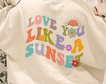 May include: A light beige sweatshirt with a colorful graphic print that says "Love You Like A Sunset". The graphic features a sunset, flowers, and a retro font.