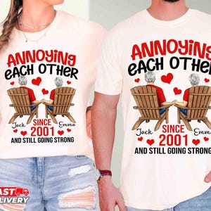 May include: Two off-white t-shirts featuring a graphic of two people sitting in wooden chairs, holding hands. The text reads: ANNOYING EACH OTHER SINCE 2001 AND STILL GOING STRONG, with the names Jack and Emma printed below the chairs and red hearts.