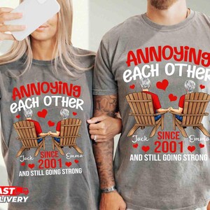 May include: A gray t-shirt featuring a graphic of two people sitting in wooden chairs, holding hands. The text reads: ANNOYING EACH OTHER SINCE 2001 AND STILL GOING STRONG, with the names Jack and Emma and several red hearts printed around the design.