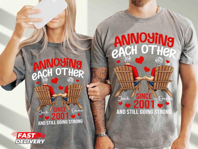 May include: A gray t-shirt featuring a graphic of two people sitting in wooden chairs, holding hands. The text reads: ANNOYING EACH OTHER SINCE 2001 AND STILL GOING STRONG, with the names Jack and Emma and several red hearts printed around the design.