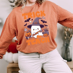 May include: Orange sweatshirt with a ghost wearing a witch hat and holding a pencil. The text "Trick or Teach" is printed on the sweatshirt.
