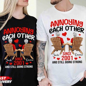 May include: A black t-shirt and a white t-shirt, each featuring a graphic of two people sitting in wooden chairs, holding hands, with red hearts and the text: ANNOYING EACH OTHER SINCE 2001 AND STILL GOING STRONG, with the names Jack and Emma.