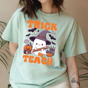 May include: A light blue t-shirt with a cute ghost wearing a witch hat and holding a pencil. The ghost is surrounded by bats, stars, and pumpkins. The text "Trick or Teach" is printed in orange on the shirt.